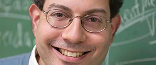 Head shot of Adam Shear, smiling man with dark hair and glasses in front of blackboard